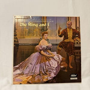 May include: A vintage vinyl record cover for the motion picture The King and I by Rodgers and Hammerstein. The cover features a person in a large, shiny lavender gown sitting next to a person in a brown and gold traditional outfit.