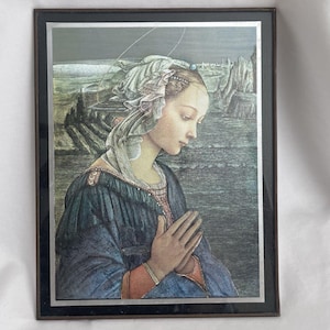May include: A framed print depicting a person with a head covering and prayerful hands, wearing a blue garment with red cuffs. The background features a landscape with rocky formations and a dark sky, all enclosed in a thin dark brown frame.