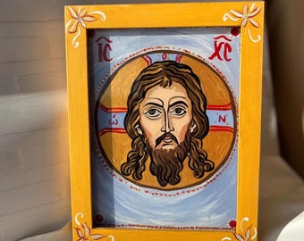 May include: A framed religious icon featuring a painted portrait of a man with long brown hair and a beard. The portrait is set against a light blue background within a circular design. The frame is yellow with floral corner accents.