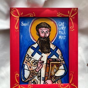 Op de afbeelding: Handgeschilderde icoon van Sint Gregorius Palamas in een rode lijst. De icoon toont een man met een donkere baard, die een boek vasthoudt, tegen een blauwe achtergrond met een gouden halo. De lijst heeft gouden decoraties.