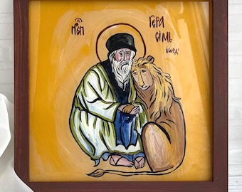 May include: A framed religious icon depicting a saint with a long white beard and a black hat, seated beside a lion. The icon is painted in warm tones of yellow, brown, and green, with text in the upper right corner. The frame is dark brown.