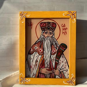 Può includere: Icona religiosa dipinta a mano in una cornice di legno gialla. L'icona raffigura una figura barbuta in una veste rossa e bianca, che tiene un bastone. La cornice ha disegni floreali agli angoli. Lo sfondo è color pesca chiaro.