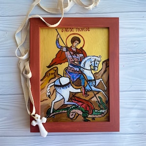 May include: A framed painting of Saint George on horseback slaying a dragon. The painting is in a red frame and has a yellow background. The text "Ο ΑΓΙΟΣ ΓΕΩΡΓΙΟΣ" is written in Greek at the top of the painting.