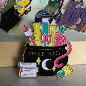 May include: Enamel pin featuring a black cauldron with the words "MAKE MAGIC". The cauldron is filled with art supplies, including a paintbrush, scissors, and yarn. Other pins in the background include books and a hedgehog.