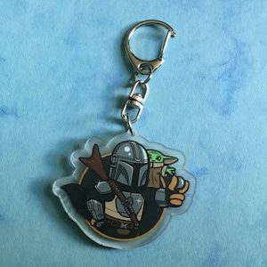 K&ouml;nnte beinhalten: Ein durchsichtiger Acryl-Schl&uuml;sselanh&auml;nger mit einer Cartoon-Illustration eines Mandalorianer-Charakters, der ein kleines gr&uuml;nes Alien h&auml;lt. Der Mandalorianer tr&auml;gt einen Helm und eine R&uuml;stung, und das Alien tr&auml;gt eine braune Robe. Der Schl&uuml;sselanh&auml;nger hat einen silbernen Metallring.