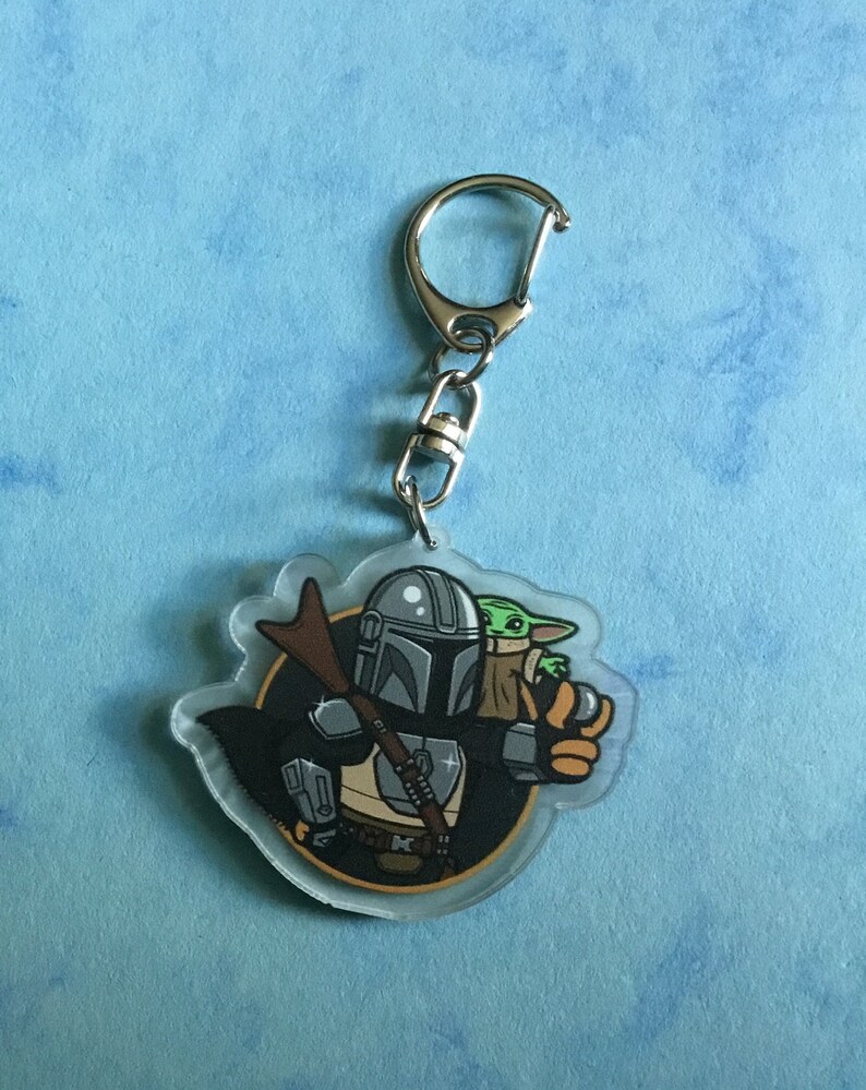 K&ouml;nnte beinhalten: Ein durchsichtiger Acryl-Schl&uuml;sselanh&auml;nger mit einer Cartoon-Illustration eines Mandalorianer-Charakters, der ein kleines gr&uuml;nes Alien h&auml;lt. Der Mandalorianer tr&auml;gt einen Helm und eine R&uuml;stung, und das Alien tr&auml;gt eine braune Robe. Der Schl&uuml;sselanh&auml;nger hat einen silbernen Metallring.