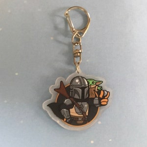 K&ouml;nnte beinhalten: Ein durchsichtiger Acryl-Schl&uuml;sselanh&auml;nger mit einer Cartoon-Illustration von Mandalorianer und Grogu. Der Mandalorianer tr&auml;gt seine charakteristische R&uuml;stung und h&auml;lt ein Gewehr. Grogu sitzt auf der Schulter des Mandalorianers und h&auml;lt ein kleines gr&uuml;nes Objekt. Der Schl&uuml;sselanh&auml;nger ist an einem silbernen Metallring befestigt.