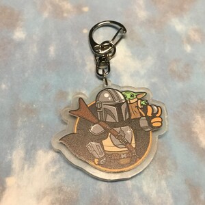 K&ouml;nnte beinhalten: Ein durchsichtiger Acryl-Schl&uuml;sselanh&auml;nger mit einer Cartoon-Illustration eines Mandalorianer-Charakters mit einem gr&uuml;nen au&szlig;erirdischen Kind in seinen Armen. Der Mandalorianer tr&auml;gt einen Helm und eine R&uuml;stung, und das Kind tr&auml;gt eine braune Robe. Der Schl&uuml;sselanh&auml;nger ist an einem silbernen Metallring befestigt.