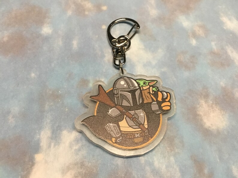 K&ouml;nnte beinhalten: Ein durchsichtiger Acryl-Schl&uuml;sselanh&auml;nger mit einer Cartoon-Illustration eines Mandalorianer-Charakters mit einem gr&uuml;nen au&szlig;erirdischen Kind in seinen Armen. Der Mandalorianer tr&auml;gt einen Helm und eine R&uuml;stung, und das Kind tr&auml;gt eine braune Robe. Der Schl&uuml;sselanh&auml;nger ist an einem silbernen Metallring befestigt.
