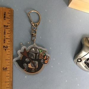 K&ouml;nnte beinhalten: Ein durchsichtiger Acryl-Schl&uuml;sselanh&auml;nger mit einer Cartoon-Illustration von Mandalorianer und Grogu aus der Star Wars-Serie. Der Mandalorianer tr&auml;gt seine charakteristische R&uuml;stung und h&auml;lt Grogu in seinen Armen. Der Schl&uuml;sselanh&auml;nger hat einen silbernen Metallring.