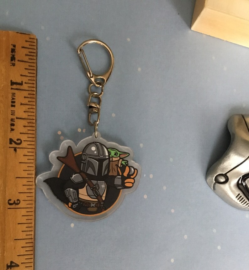K&ouml;nnte beinhalten: Ein durchsichtiger Acryl-Schl&uuml;sselanh&auml;nger mit einer Cartoon-Illustration von Mandalorianer und Grogu aus der Star Wars-Serie. Der Mandalorianer tr&auml;gt seine charakteristische R&uuml;stung und h&auml;lt Grogu in seinen Armen. Der Schl&uuml;sselanh&auml;nger hat einen silbernen Metallring.
