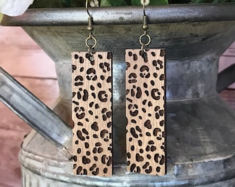 Op de afbeelding: Een paar houten oorbellen met een luipaardprint. De oorbellen zijn bruin met een lichtbruine luipaardprint. De oorbellen hangen aan gouden haakjes.