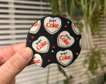 May include: A black and white coaster with a repeating pattern of white hearts with red "DIET COKE" text. The hearts are outlined in black and have a white background.