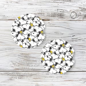 May include: Two round car coasters with a repeating pattern of Snoopy's face in black and white, with yellow stars. The coasters are on a light wood-grain surface.