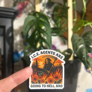 May include: A sticker with a grim reaper graphic, flames, and text that reads "I.C.E. AGENTS ARE GOING TO HELL, BRO." The sticker has a white border and is held in front of a potted plant.
