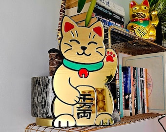 May include: A gold-colored, smiling Maneki-neko cat figurine with a raised paw, green collar, and red accents. The cat holds a sign with Japanese characters. The figurine sits on a gold wire shelf, with a smaller gold cat figurine and books in the background.