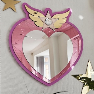 Puede incluir: Un espejo en forma de corazón con marco rosa y superficie espejada. El espejo está adornado con alas doradas y un adorno en forma de estrella con un cristal central. También hay dos decoraciones doradas en forma de estrella.