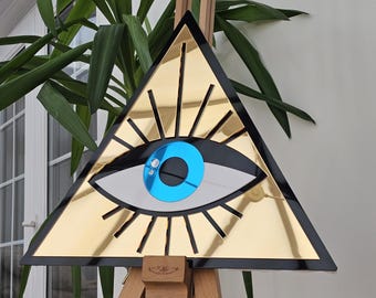 Könnte beinhalten: Ein goldfarbenes, dreieckiges Wandkunstwerk mit einem stilisierten Augendesign. Das Auge hat eine blaue Iris, ein silberfarbenes Auge und schwarze Akzente. Das Kunstwerk ist auf einer hölzernen Staffelei montiert, mit grünen Pflanzen im Hintergrund.