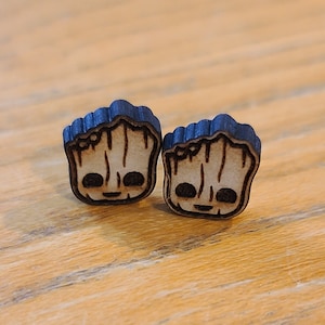 May include: Two wooden earrings featuring a cartoon image of a character from the Guardians of the Galaxy movie. The character is a tree-like humanoid with a brown face and black eyes. The earrings are painted with black details and have a dark brown wood grain.