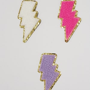 Puede incluir: Tres parches termoadhesivos en forma de rayo en blanco, rosa y morado con ribete de purpurina dorada. Cada parche mide 7 cm por 4 cm.