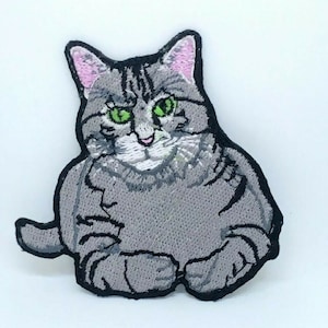 May include: Iron-on patch featuring an embroidered grey tabby cat with green eyes and pink ears. The cat is sitting with its paws tucked under its body.