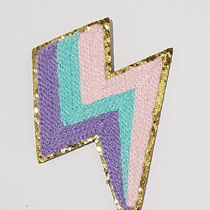 Puede incluir: Un parche con forma de rayo de colores con un contorno de purpurina dorada. El parche está hecho de hilo rosa, morado y turquesa. El parche mide 8,5 x 5 cm.