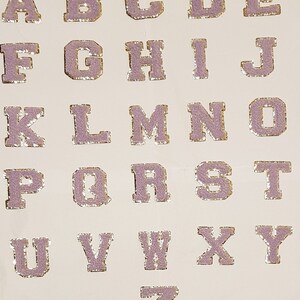 May include: A complete alphabet of lavender sequined letters with gold edging, displayed on a white surface. Each letter is individually cut. The letters are in a classic serif font, suitable for various craft projects.