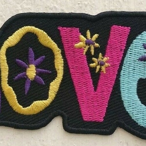 Op de afbeelding: Gestikt patch met het woord "LOVE" in kleurrijke, gestileerde letters. De letters zijn omrand met geel en versierd met paarse bloemen.