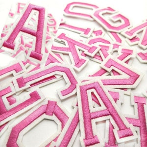 May include: A pile of pink felt letters with white outlines. The letters are all uppercase and are arranged in a random order.