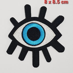 May include: A blue eye with white and black accents, surrounded by black rays. The eye is embroidered on a black fabric patch, measuring 8 x 8.5 cm.