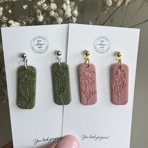 May include: Two pairs of dangle earrings with a floral design. One pair has green clay earrings with silver hooks and the other pair has pink clay earrings with gold hooks. The cards they are on say "You look gorgeous!"