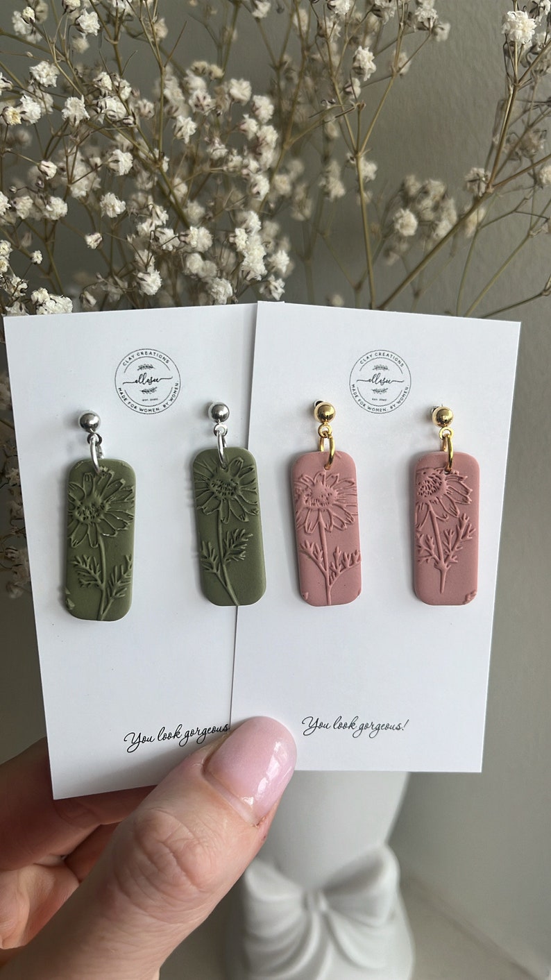 May include: Two pairs of dangle earrings with a floral design. One pair has green clay earrings with silver hooks and the other pair has pink clay earrings with gold hooks. The cards they are on say "You look gorgeous!"