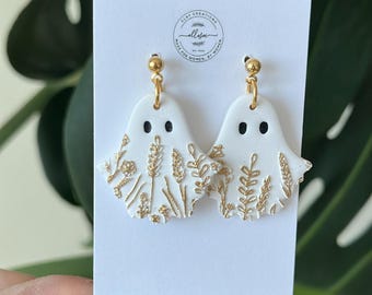May include: White ghost-shaped dangle earrings with gold leaf details. The earrings have gold hooks.