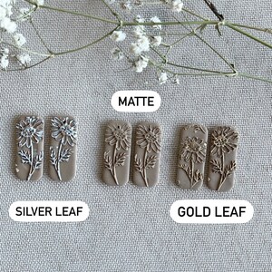 May include: Three pairs of rectangular ceramic earrings with a floral design. The earrings are in three finishes: matte, silver leaf, and gold leaf.