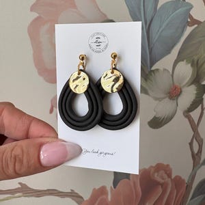 Op de afbeelding: Zwarte druppelvormige oorbellen met goudkleurige cirkelvormige accenten. De oorbellen zijn gemaakt van een donker materiaal en hebben een gelaagd ontwerp. De tekst "You look gorgeous!" staat op de witte kaart.