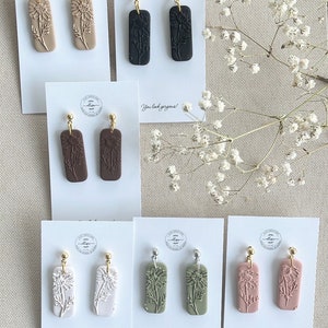 May include: Assortment of eight pairs of handmade polymer clay earrings. Each pair features a different colour and design of a flower with a gold or silver hook. The cards have the text "You look gorgeous!" printed on them.