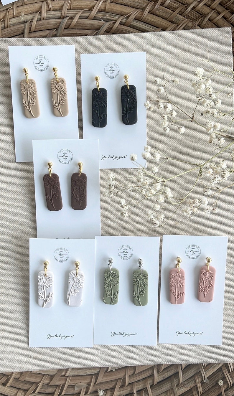 May include: Assortment of eight pairs of handmade polymer clay earrings. Each pair features a different colour and design of a flower with a gold or silver hook. The cards have the text "You look gorgeous!" printed on them.