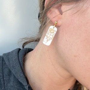 May include: A pair of white rectangular earrings with a gold floral design. The earrings are clip-on style.