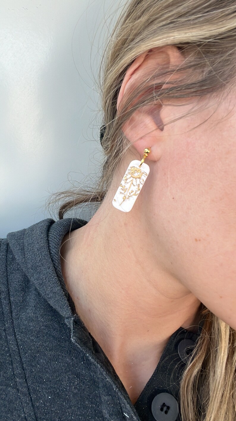 May include: A pair of white rectangular earrings with a gold floral design. The earrings are clip-on style.