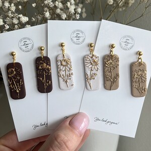 May include: Four pairs of dangle earrings with gold accents. Each pair features a rectangular shape with a different colour background: brown, white, beige, and light brown. The earrings have a gold flower design with a gold wire loop.