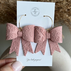 May include: A pair of pink bow earrings with a floral pattern. The earrings are made of polymer clay and have gold hoops. The earrings are displayed on a white card with the text "You look gorgeous!"