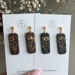 May include: Two pairs of dangle earrings with gold accents. The earrings feature a rectangular shape with a floral design in gold on a brown and black background. The text "You look gorgeous!" is printed on the white card behind the earrings.