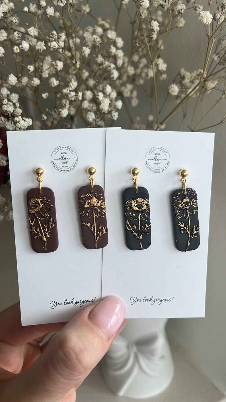May include: Two pairs of dangle earrings with gold accents. The earrings feature a rectangular shape with a floral design in gold on a brown and black background. The text "You look gorgeous!" is printed on the white card behind the earrings.