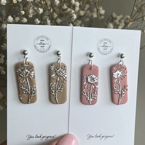 May include: Two pairs of dangle earrings with silver flower designs. One pair is on a light brown background and the other pair is on a light pink background. The earrings are handmade and have the text "You look gorgeous!" on the cards.
