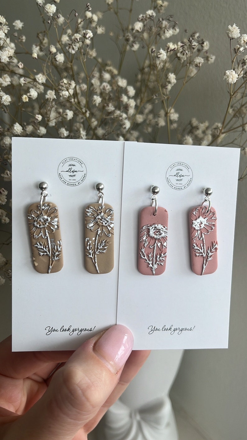 May include: Two pairs of dangle earrings with silver flower designs. One pair is on a light brown background and the other pair is on a light pink background. The earrings are handmade and have the text "You look gorgeous!" on the cards.