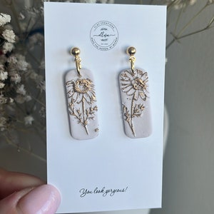 May include: A pair of white rectangular dangle earrings with gold accents. The earrings feature a gold daisy design. The earrings are displayed on a white card with the text "You look gorgeous!"