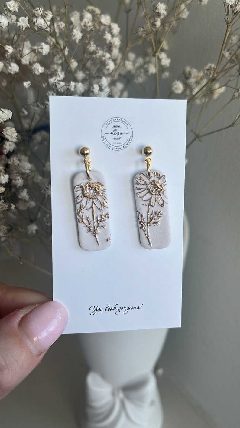 May include: A pair of white rectangular dangle earrings with gold accents. The earrings feature a gold daisy design. The earrings are displayed on a white card with the text "You look gorgeous!"