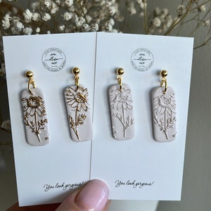 May include: A pair of white clay earrings with gold accents featuring a floral design. The earrings are displayed on a white card with the text "You look gorgeous!" printed on it.