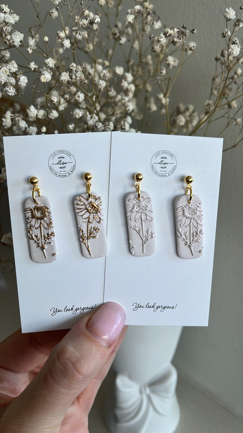 May include: A pair of white clay earrings with gold accents featuring a floral design. The earrings are displayed on a white card with the text "You look gorgeous!" printed on it.
