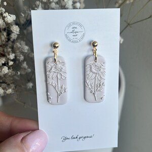 May include: A pair of white clay earrings with a floral design. The earrings are rectangular in shape and have a gold hook.